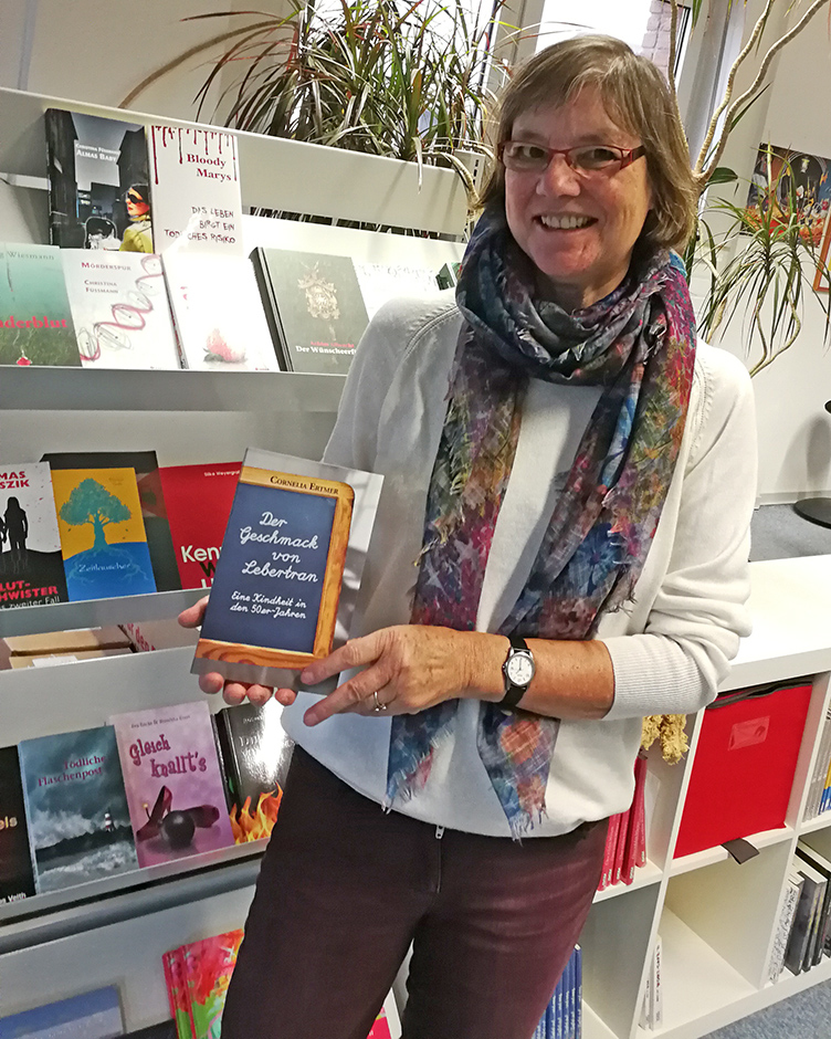 Cornelia Ertmer steht vor einem Bücherregal im Verlag und hält ihr Buch in den Händen.