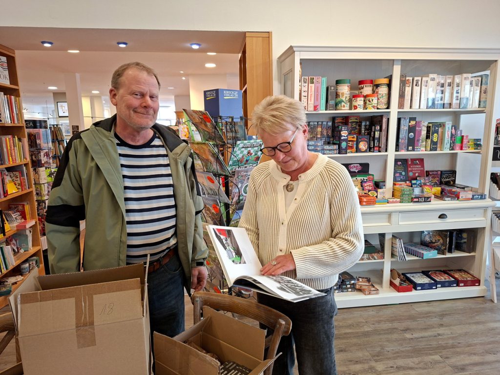Eine Frau mit Brille und hellblau-gestreifter Strickjacke durchblättert in einer Buchhandlung aufmerksam ein Exemplar des Fotobands „Zweimal Haltern am See“. Neben ihr steht ein Mann in einer grün-schwarzen Jacke und blickt lächelnd in die Kamera. Im Vordergrund stehen geöffnete Kartons.