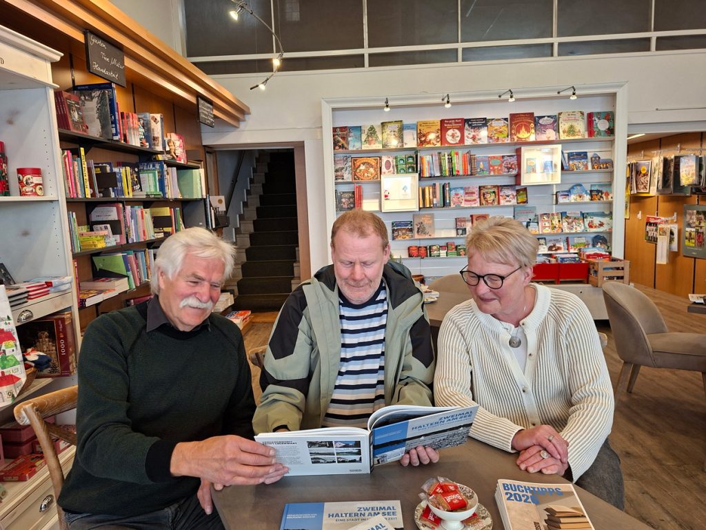 Drei Personen sitzen an einem Tisch in einer Buchhandlung und schauen gemeinsam mit Interesse in ein geöffnetes Exemplar des Buchs „Zweimal Haltern am See“. Auf dem Tisch liegen weitere Buchtipps-Broschüren und eine kleine Schale mit Keksen.