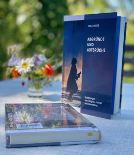 Das Buch „Abgründe und Aufbrüche“ von Enka Wiese steht aufrecht auf einem hellen Gartentisch, daneben liegt ein zweites Exemplar und ein kleines Glas mit bunten Wiesenblumen vor grünem Hintergrund.