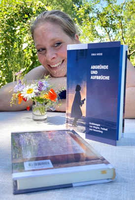 Enka Wiese lehnt entspannt im Hintergrund und lächelt, während im Vordergrund ihr Buch „Abgründe und Aufbrüche“ auf einem Gartentisch präsentiert wird.