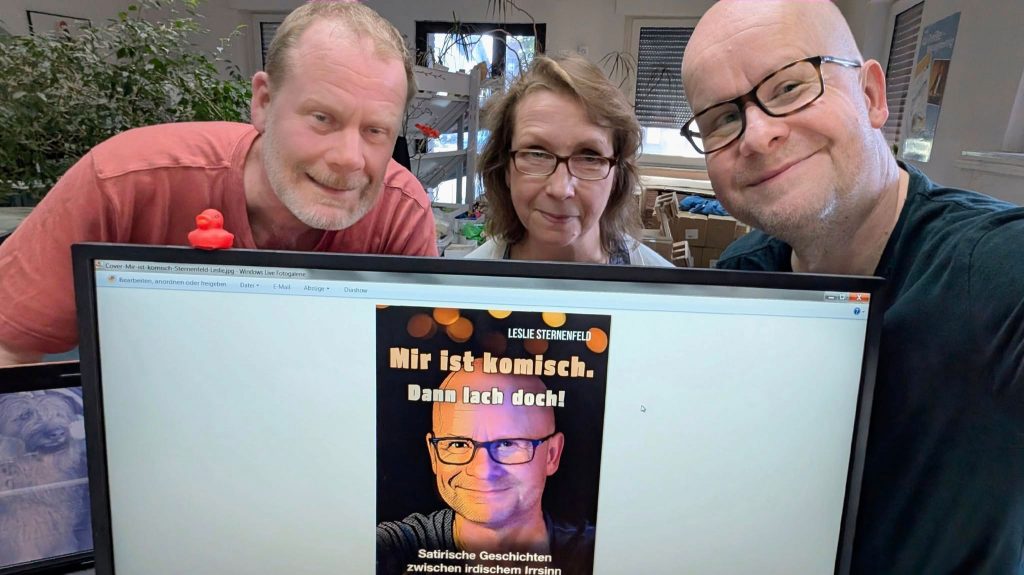 Leslie Sternenfeld mit den Verlegern Elke Neumann und Georg Nies hinter einem Monitor. Im Monitor ist das Cover von Leslies neuem Buch zu sehen.