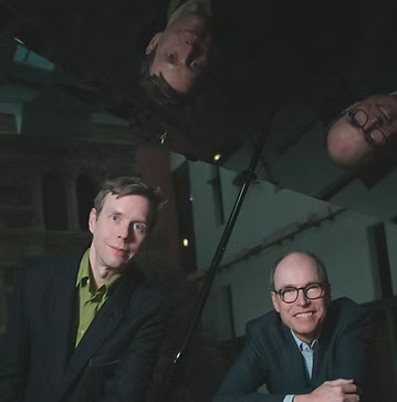 Martin Brödemann (links) und Thomas Eicher (rechts), beide in Anzügen, blicken lächelnd in die Kamera. Ihr Bild spiegelt sich kopfüber im geöffneten, glänzenden Deckel eines Flügelpianos.