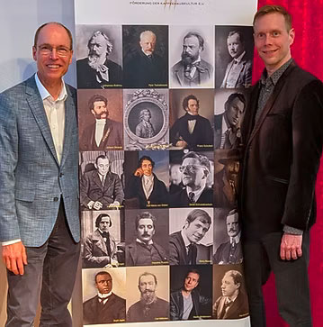 Thomas Eicher (links) und Martin Brödemann (rechts) stehen neben einem großen Roll-up-Banner, das eine Galeriesammlung historischer Porträts klassischer Komponisten zeigt.