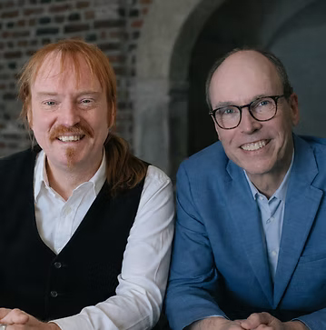 Carsten Bülow (links) und Thomas Eicher (rechts) sitzen nebeneinander in einem Innenraum und lächeln in die Kamera. Carsten Bülow hat langes rotes, zurückgebundenes Haar, einen roten Schnurrbart und Kinnbart und trägt ein weißes Hemd mit schwarzer Weste. Thomas Eicher hat kurze Haare, trägt eine Brille sowie einen blauen Blazer über einem hellblauen Hemd. Im Hintergrund ist eine Backsteinwand mit einem gewölbten Türbogen zu sehen.